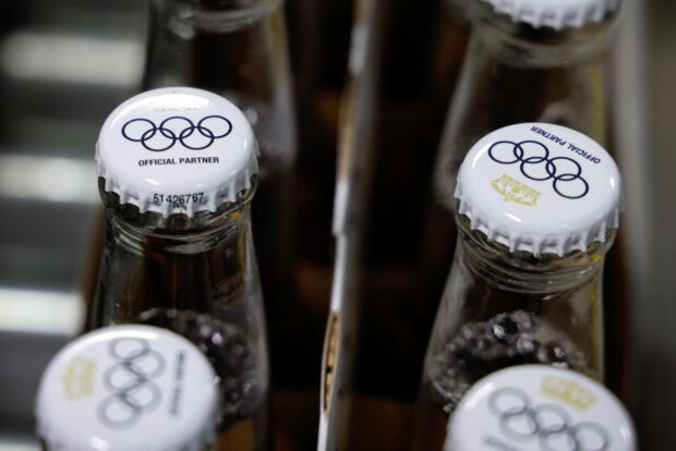 This photograph taken on April 23, 2024 shows crown caps covering beer bottles filled with Olympics logo on bottles of Corona Cero alcohol-free beer on a production line during a press visit at the Anheuser-Busch InBev (AB InBEV) brewery in Leuven. AB InBev, will be a World Olympic Partner until 2028. Corona Cero alcohol-free beer thus becomes a global sponsor of Olympic Games. (Photo by Kenzo TRIBOUILLARD / AFP)