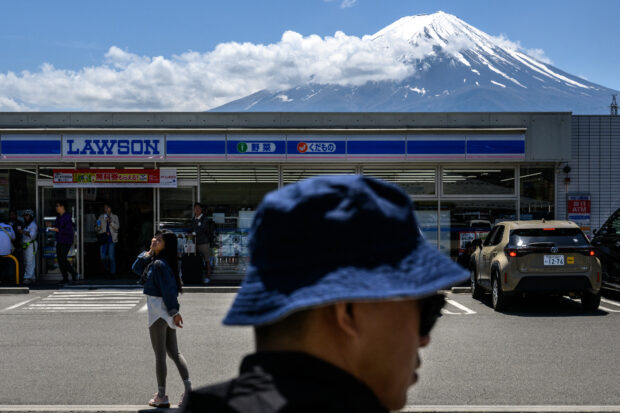 A tourist poses in front of a convenience store with Mount Fuji on May 3, 2024, before a huge black barrier which will be installed to block Mount Fuji from view, in the town of Fujikawaguchiko, Yamanashi prefecture.